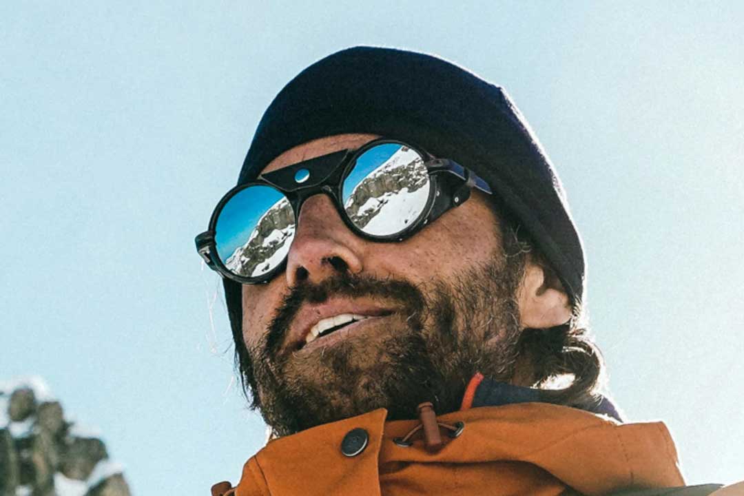 Upward view of man wearing round glacier sunglasses frame with side shields and orange jacket in mountains on a bright sunny day