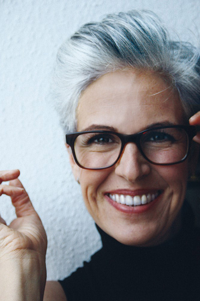 Woman smiling with grey combed hair wearing rounded eyeglasses frame