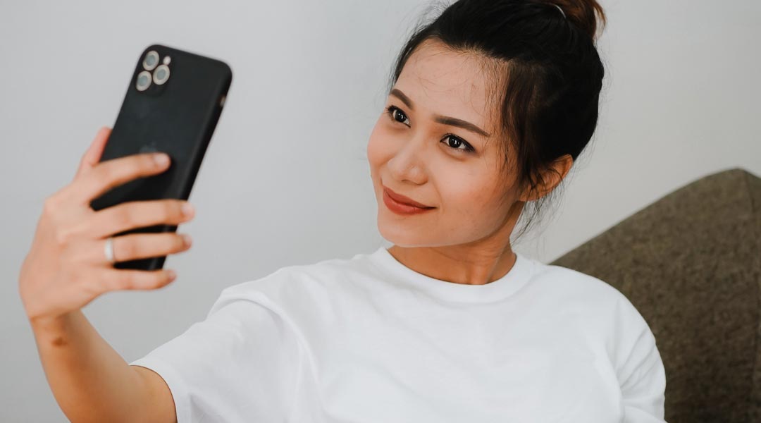 Woman taking a video of herself on a mobile phone smiling wearing a white T shirt sitting on a sofa