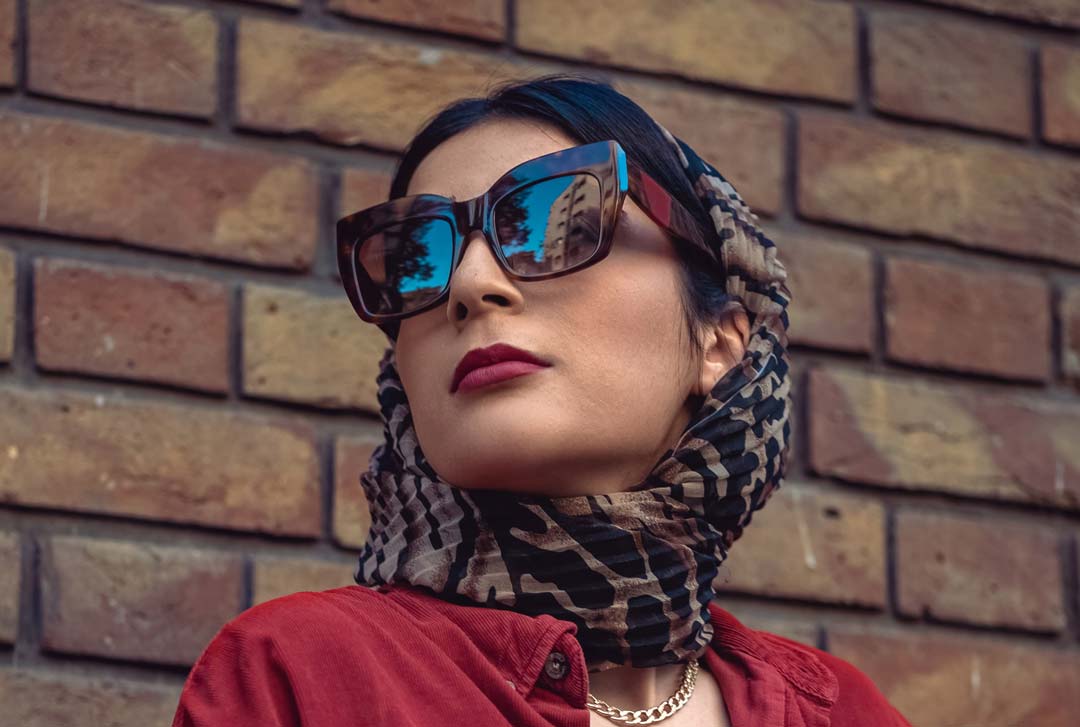 Woman wearing large square sunglasses frame and head scarf in front of brick wall looking to her side