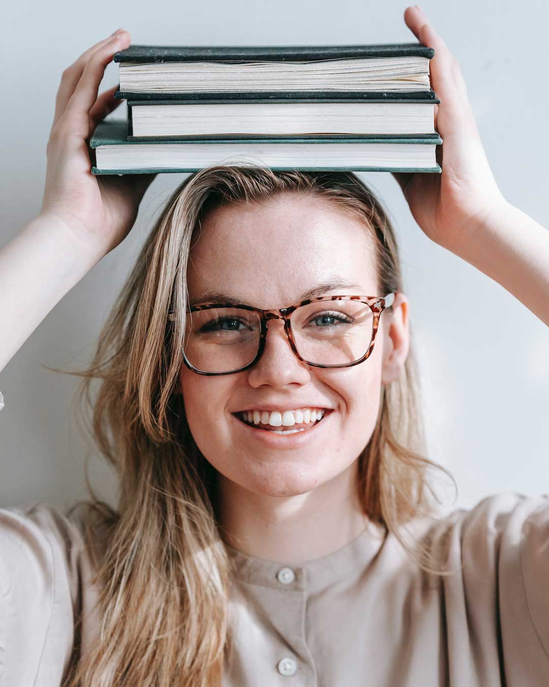 Woman with blonde hair wearing oversized tortoise eyeglasses holding books above her head