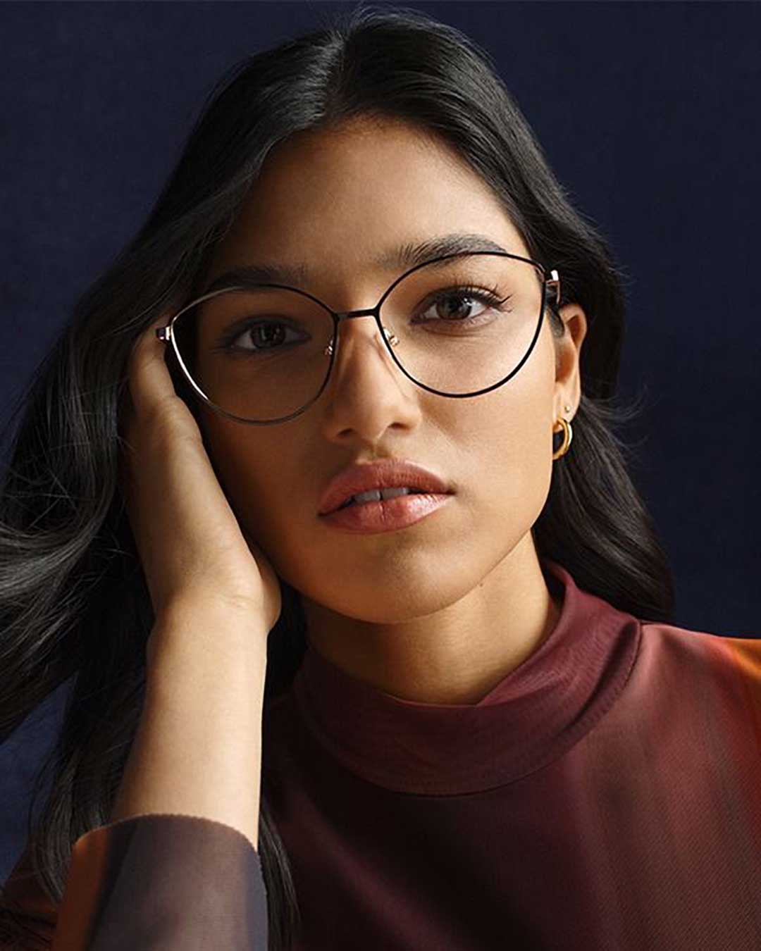 Woman with long dark hair wearing cat eye glasses frame in front of blue background