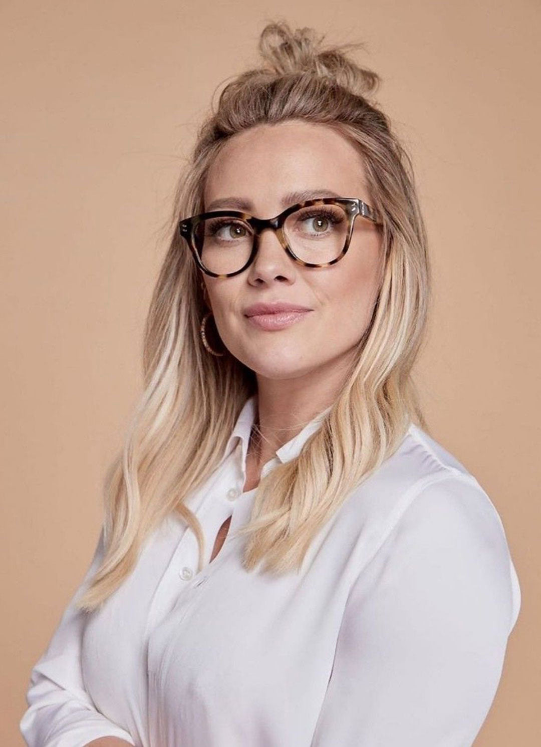 Woman with tied up blonde hair in front of beige background wearing white shirt and tortoise glasses