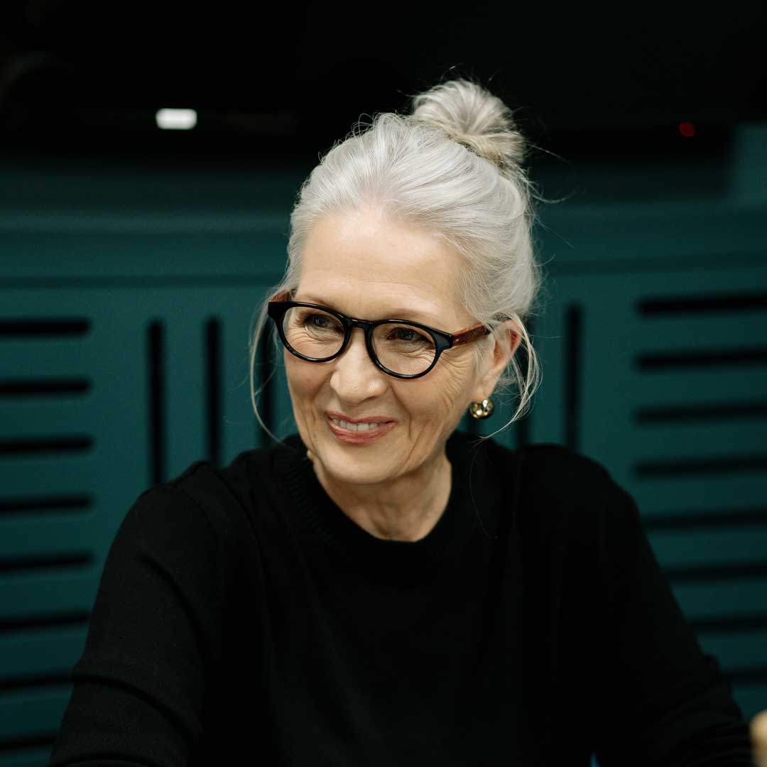 Woman with tied up grey hair sitting at desk smiling wearing oval black eyeglasses