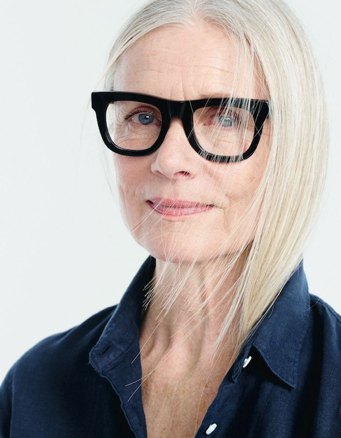 Woman with long grey hair and blue shirt wearing a very thick black glasses frame