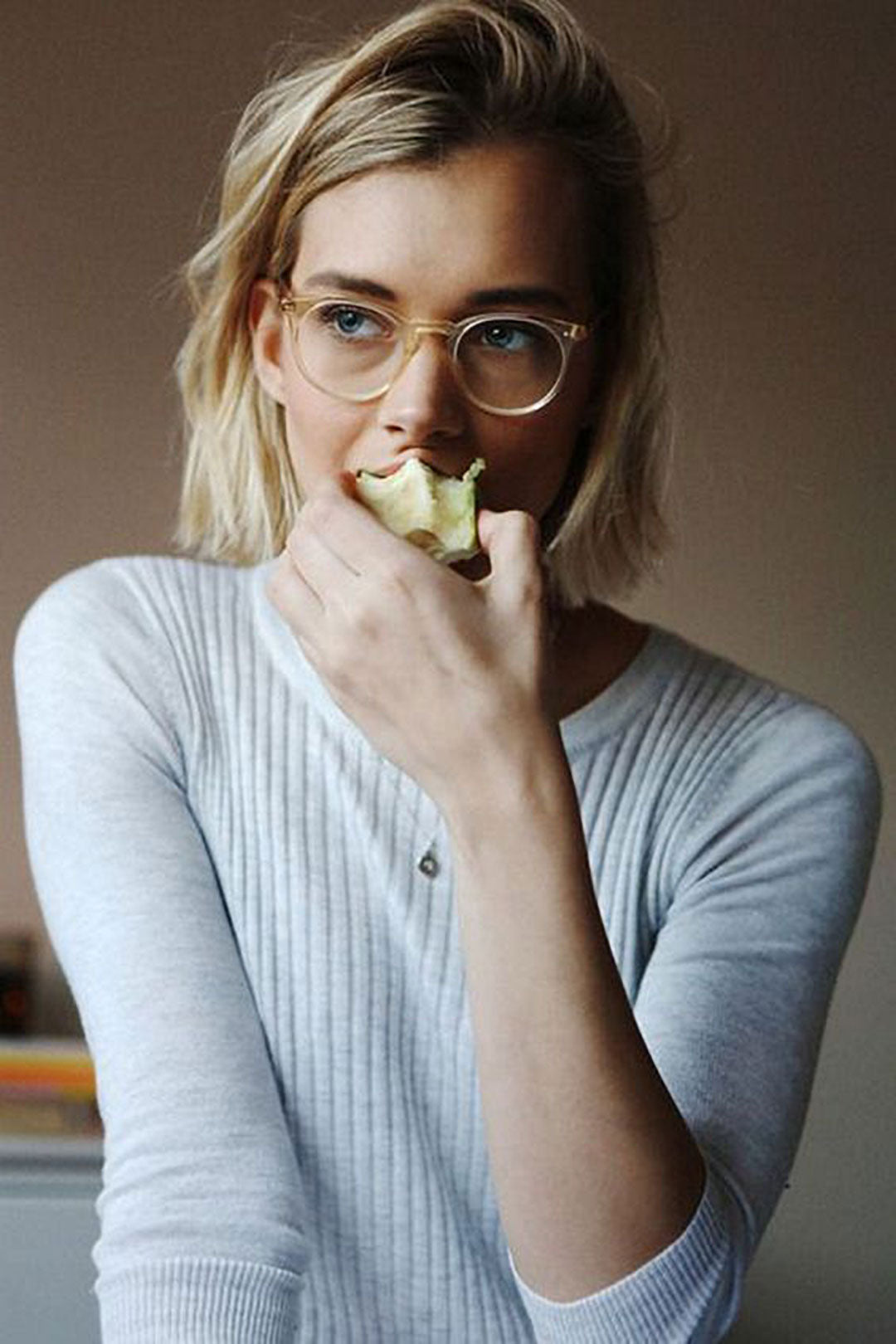 Young blonde female wearing clear glasses frame and grey jumper eating an apple
