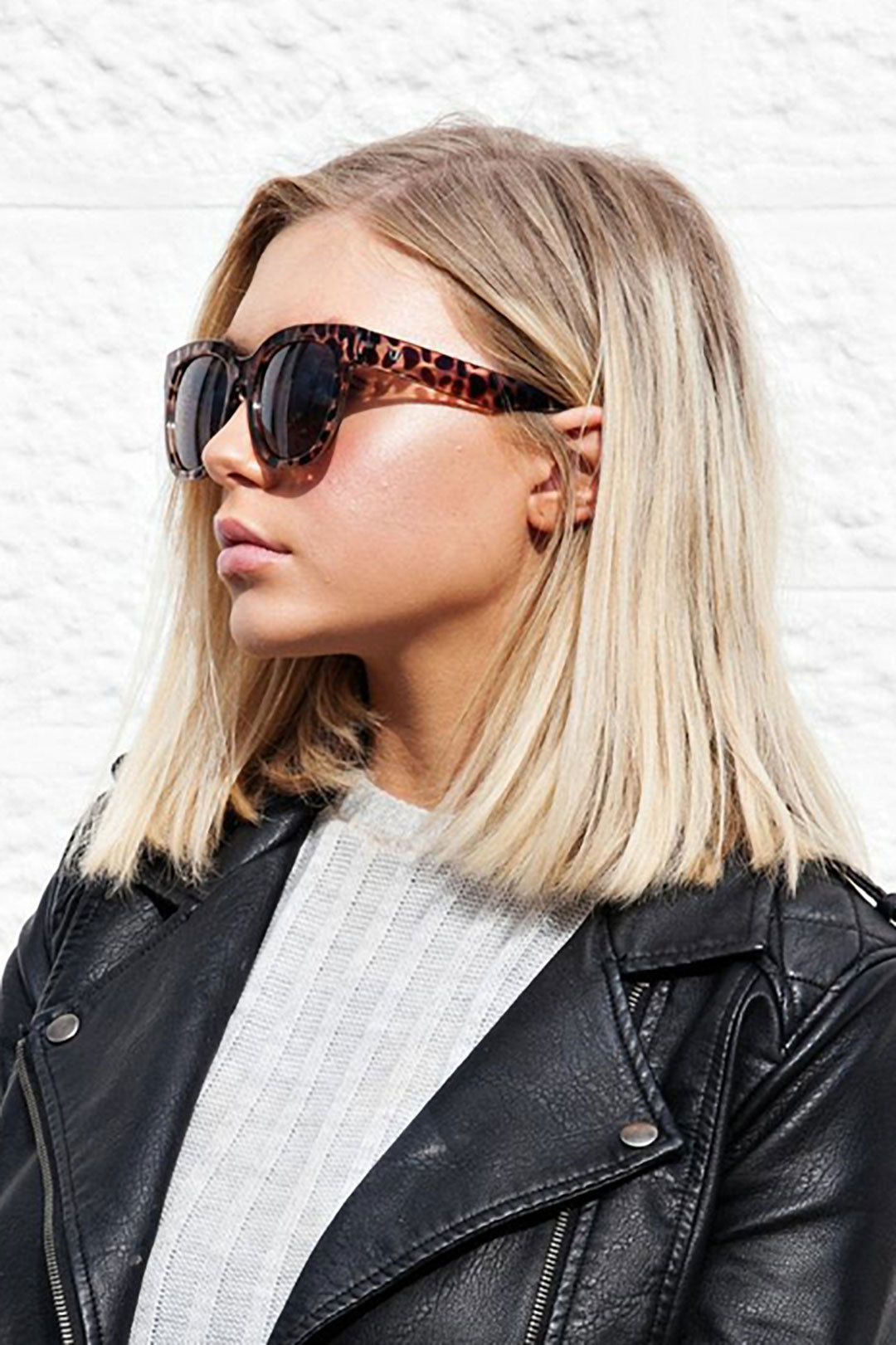 Young blonde lady in front of white wall wearing black leather jacket and large tortoiseshell sunglasses