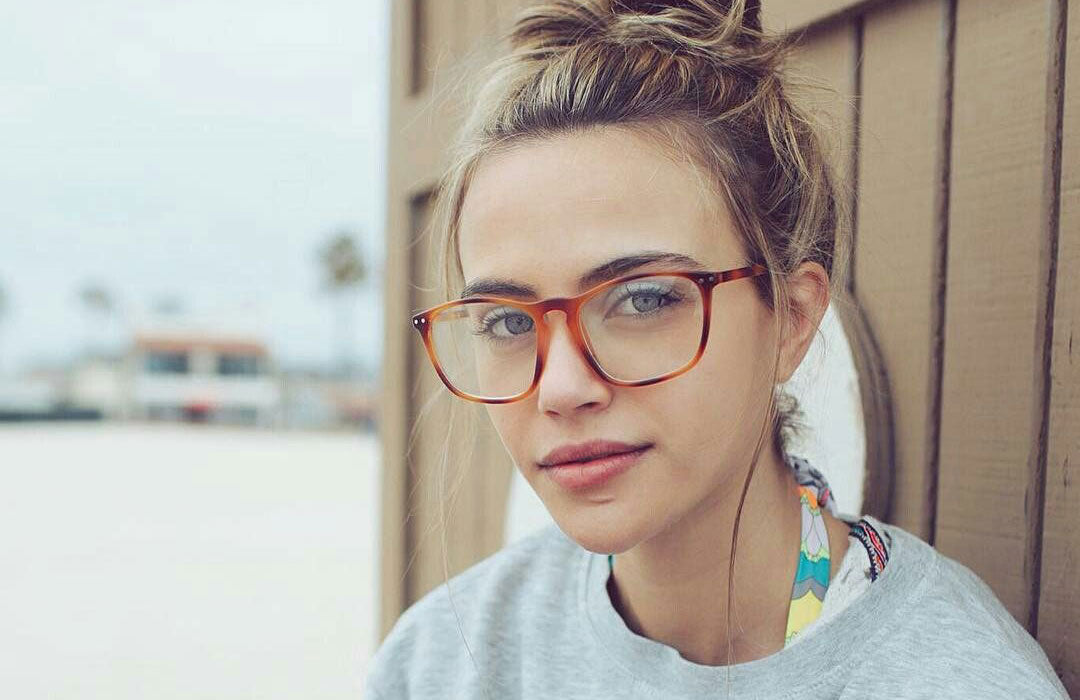 Young blonde lady sitting at beach hut wearing grey jumper and amber eyeglasses
