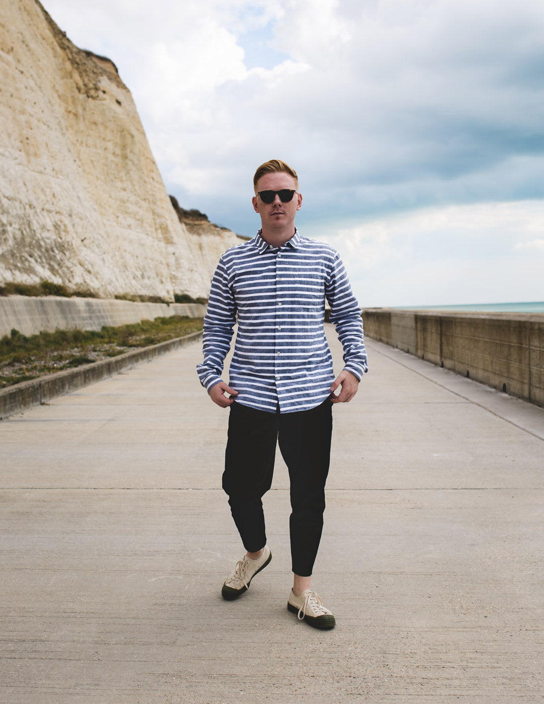 Young male walking beside beach wearing stripey blue shirt and tortoise sunglasses