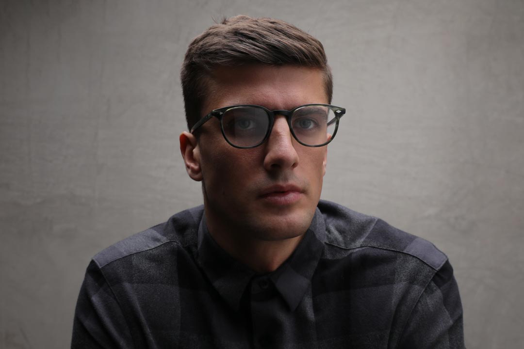 Young man with brown hair wearing oval green spectacles and dark checked shirt