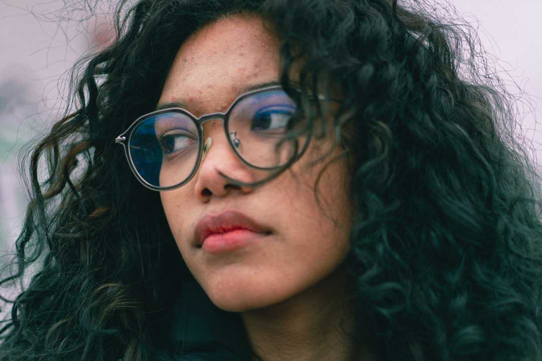 Young woman with curly black hair wearing black rimmed spectacles looking away