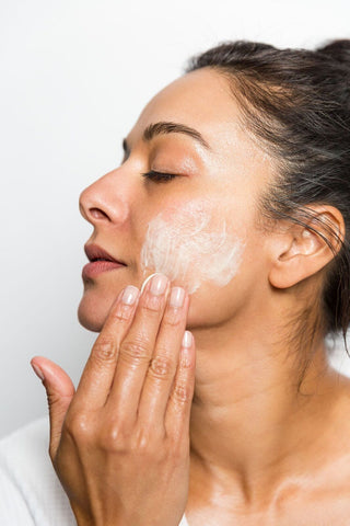 woman with eyes closed applying white skin cream to face