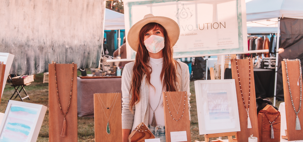 The founder of Mala Revolution stands in her pop-up shop in Venice, California where her handmade bracelets and handmade malas are displayed.  Her delicate bracelets and delicate necklaces are arranged in front of her and she smiles at the camera while wearing a mask made with original artwork. 