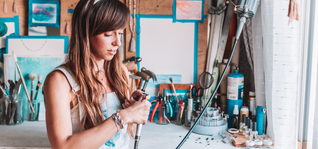 The founder of Mala Revolution in her art studio working on handmade bracelets and handmade malas and handmade jewelry.  She holds a handcrafted sterling silver charm with a lotus flower. 