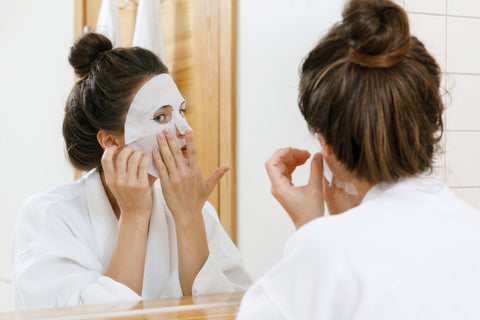 woman looking in a mirror while applying a sheet mask