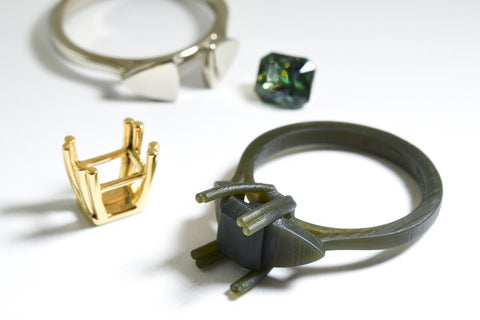 A green wax model of a gemstone ring, and a white gold shank and a yellow gold component, along with a green sapphire gemstone: the parts of a ring that will be hand made.