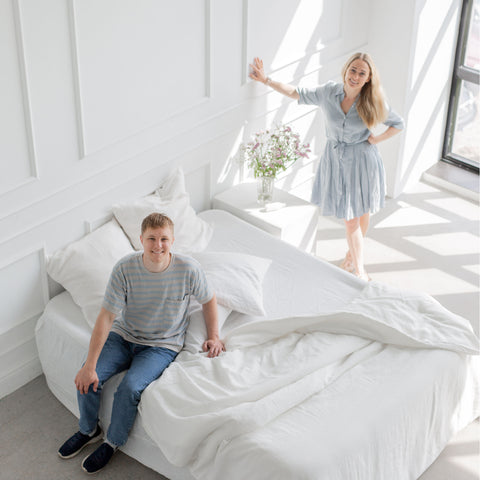 A young couple poses around a brilliant white linen bed. They look happy and confident.