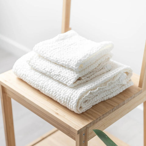 A stack of Linen & letters white linen waffle weave towels. Face cloth, on a hand towel, one a bath towel - placed on a bamboo chair.