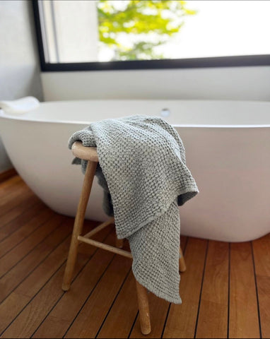 A Linen & Letters sage green waffle linen towel, draped on a wooden stool next to a sleek modern bathtub.