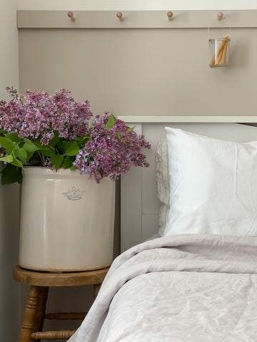 Linen and Letters soft lavendar linen duvet cover, with white linen pillowcases. Lavender blooms sit in a vintage ceramic jug on the side table. 