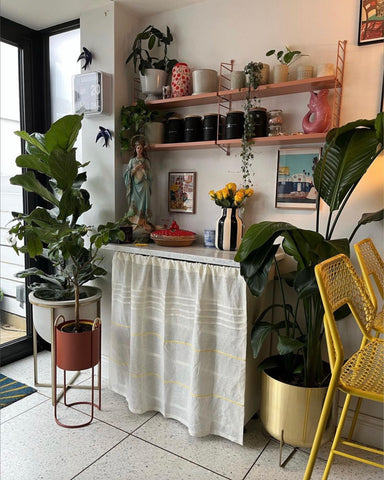 Eclectic sideboard decorated with accessories in bold colours. The front of the sideboard is covered in linen curtain with decorative yellow embroidery that matches the bold yellow wire breakfast stool.