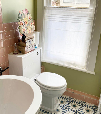 A small bathroom with a dramatic floral tile floor. Painted a bold green paired with coral wall tiles, the white linen curtain brightens the room.