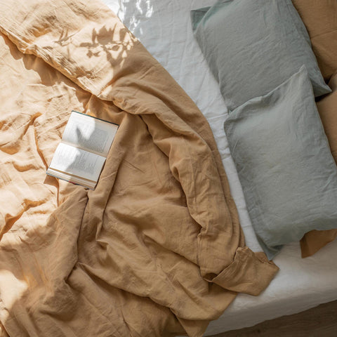 A comfy rumpled bed with an ocher linen duvet cover and sage linen pillowcases. An open book lies on top.