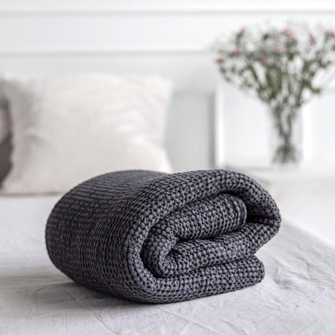 A rolled up charcoal linen waffle-weave blanket sits on a pristine white linen bed.