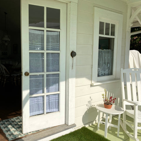 A photo of a beach house entrance. The glass panelled door stands invitingly open and features a lovely linen door curtain behind the glass.