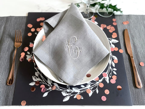 A grey linen napkin makes a placesetting centrepiece. Embroidered with a shiny grey monogram, the napkin sits on a white textured plate surrounded by large pink confetti.