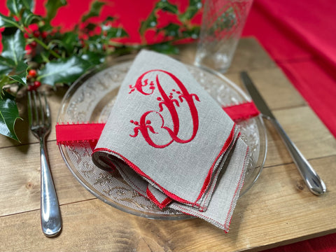 A natural linen napkin is folded and set upon a plate on a rustic wood table. The napkin is embroidered with a large red letter D and the edges are finished with a wavy red stitch.