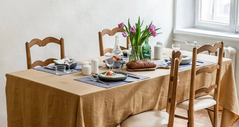 A country table is dressed with an ocher linen table cloth and a vase of flowers, ready for a spring family meal.