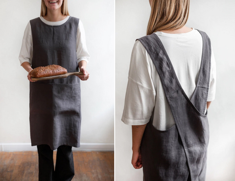 A woman wearing a dark charcoal grey apron carrying a loaf of bread on a wooden board. A woman facing away to show the crossed straps at the back of the pinafore apron.