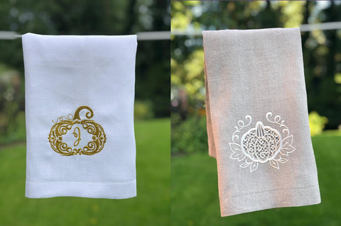 Photo of a white linen tea towel embroidered with a gold pumpkin monogram and a natural linen tea towel embroidered with a white cutwork pumpkin. Both linen towels are hanging from a washing line.