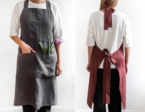 A man wearing a charcoal dark grey apron with purple flowers in the pocket. A woman stood with her back to you, wearing a terracotta red apron with lovely bows tided at the neck and back.