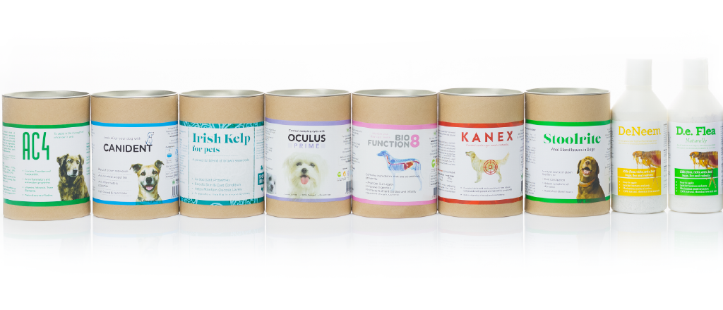 A lineup of seven seaweed based pet care products on a reflective white surface. From left to right: 'AC4', a green-labeled container with a dog image; 'CANIDENT', with a blue and white label showing a smiling dog; 'Irish Kelp for Pets', in blue with a picture of a dog and cat; 'OCULUS PRIME', in purple, featuring an image of a dog's eye; 'BIOFUNCTION8', in pink, with various pet images; 'KANEX', in green, showing a dog's digestive system; and two spray bottles labeled 'DeNeem' and 'De.Flea Naturally', both with images of dogs and claims of natural ingredients for flea control.