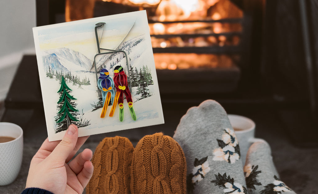 A hand holding the Quilled Ski Lift Greeting Card while sitting in front of an open fire. 