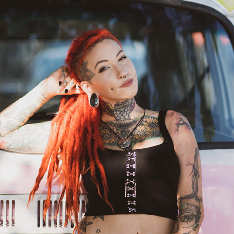 Woman with dreadlocks and tattoos standing in front of car with one hand on head