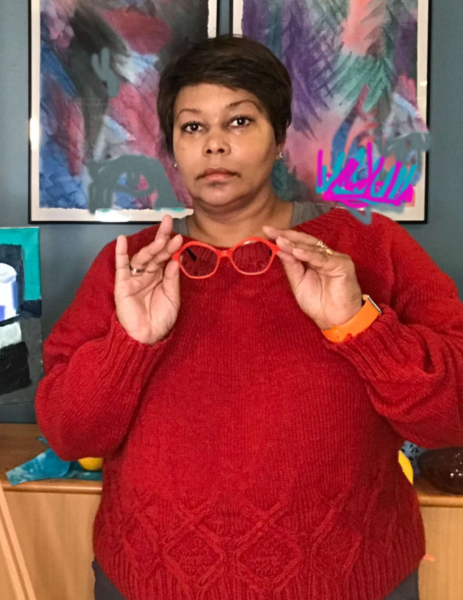 an overweight, middle-aged Black/African-American woman wearing a red cabled sweater holding a pair of red glasses