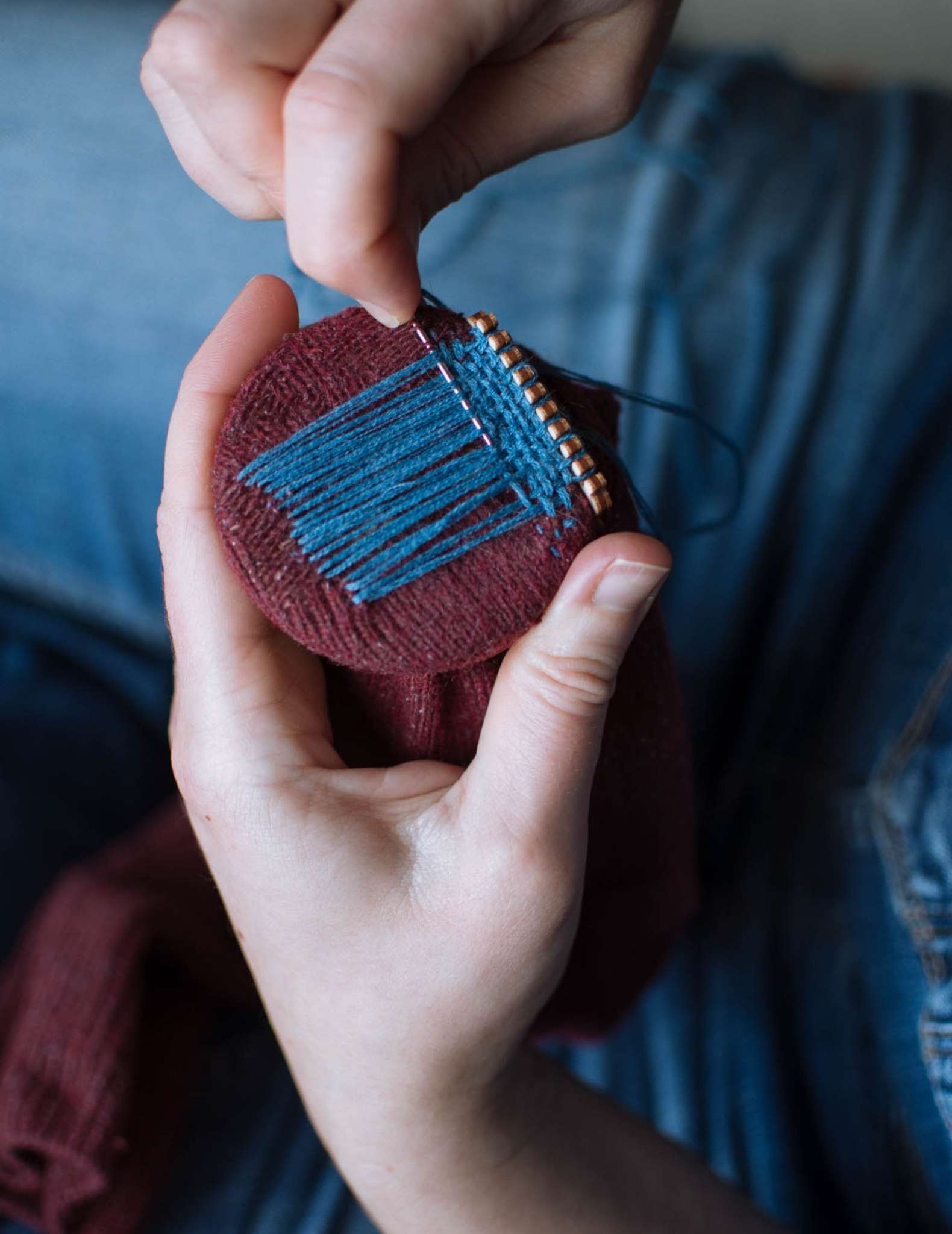 A model holds a burgundy sock while darning it with teal blue thread, using a wooden darning loom.