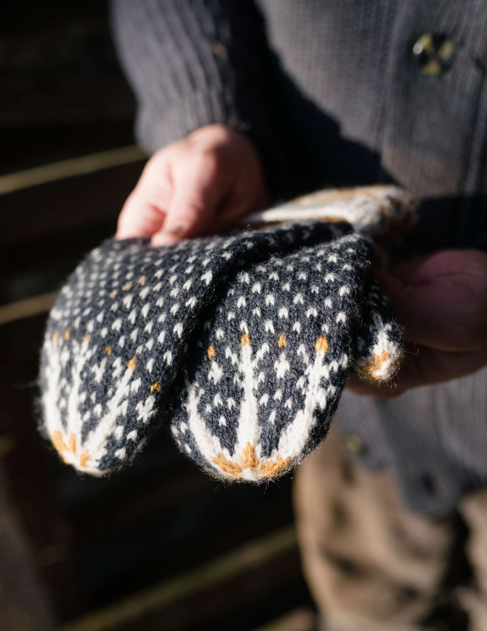 A pair of colourwork mittens in dark blue, white and yellow are held flat, unworn by a pair of white hands outdoors.