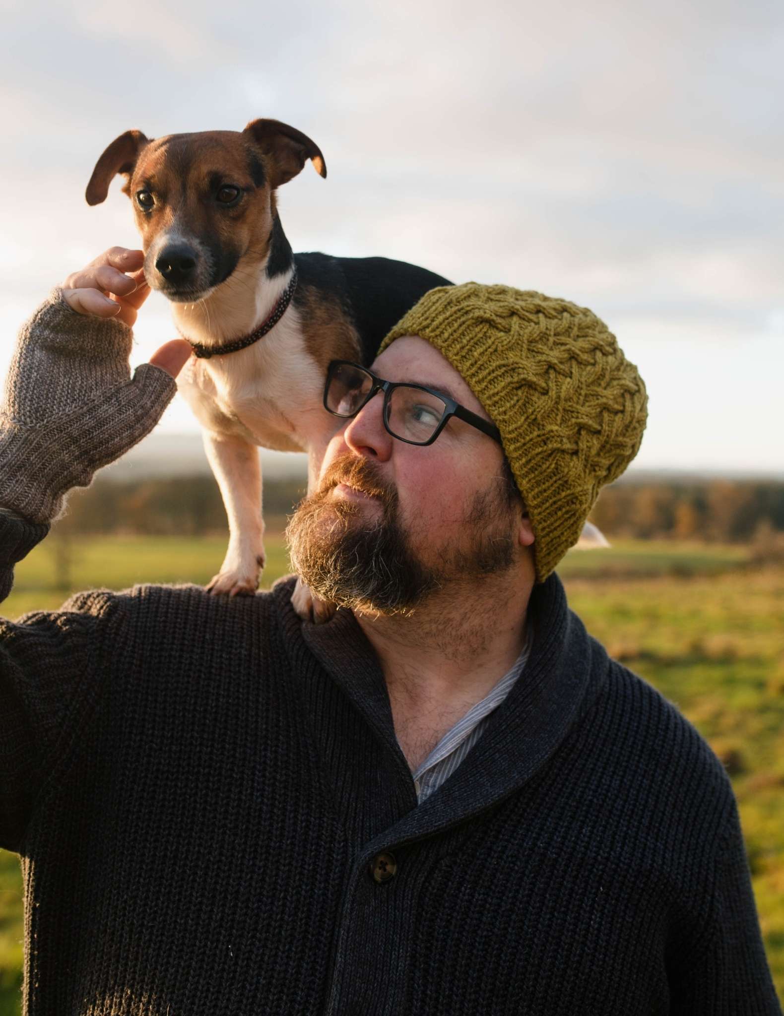 A white man with a beard and glasses stands in a field wearing a cabled green beanie hat. There is a small brown, white and dark dog standing across his shoulders and he has his hand raised towards it.
