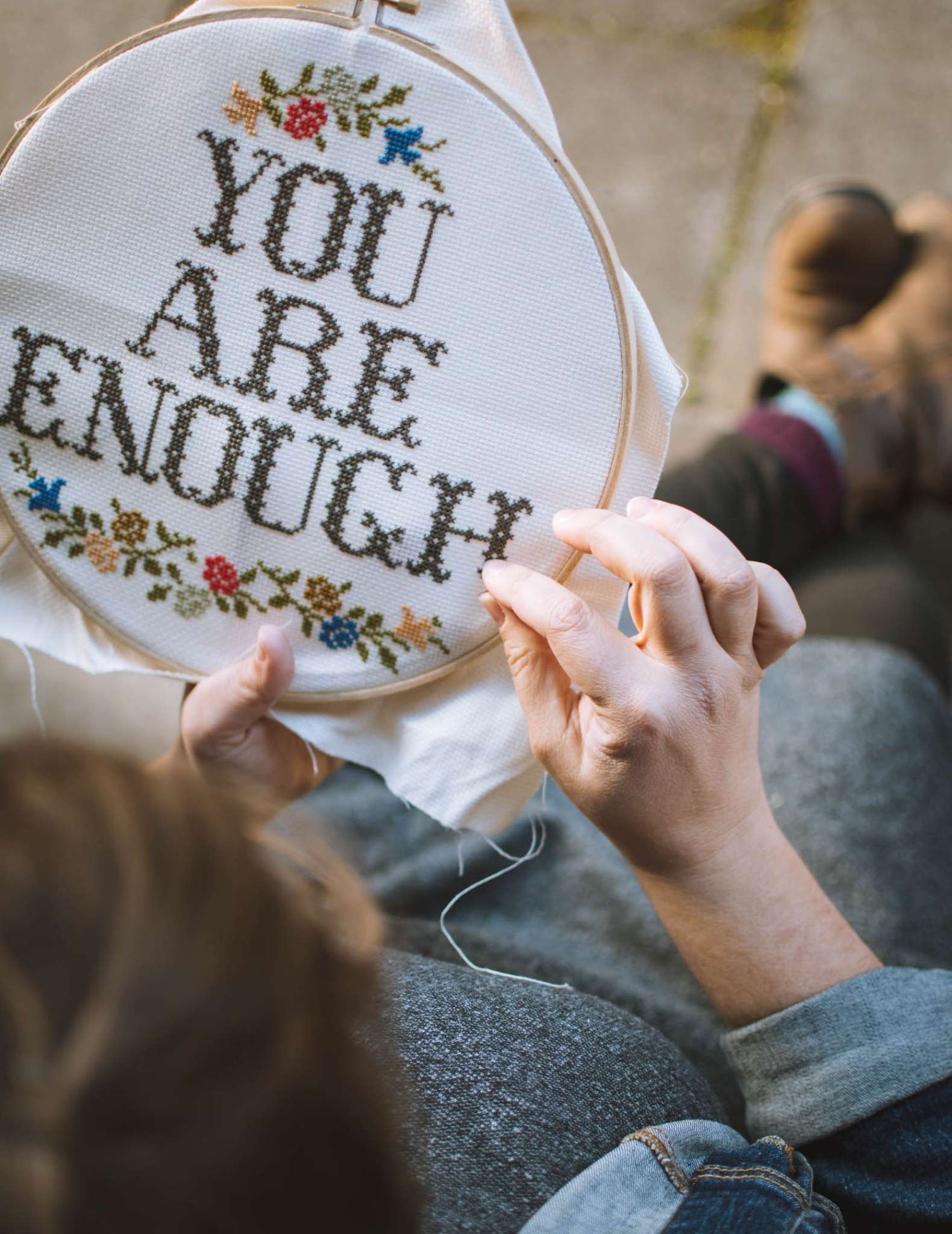 A white model with brown hair embroiders a cross stitch piece in a hoop showing the text 'You are Enough'