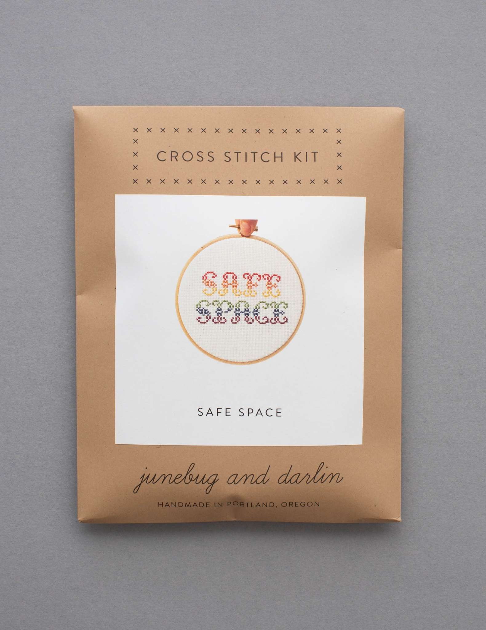 A cross stitch kit in a brown envelope showing a design in rainbow colours with the text 'Safe Space.'