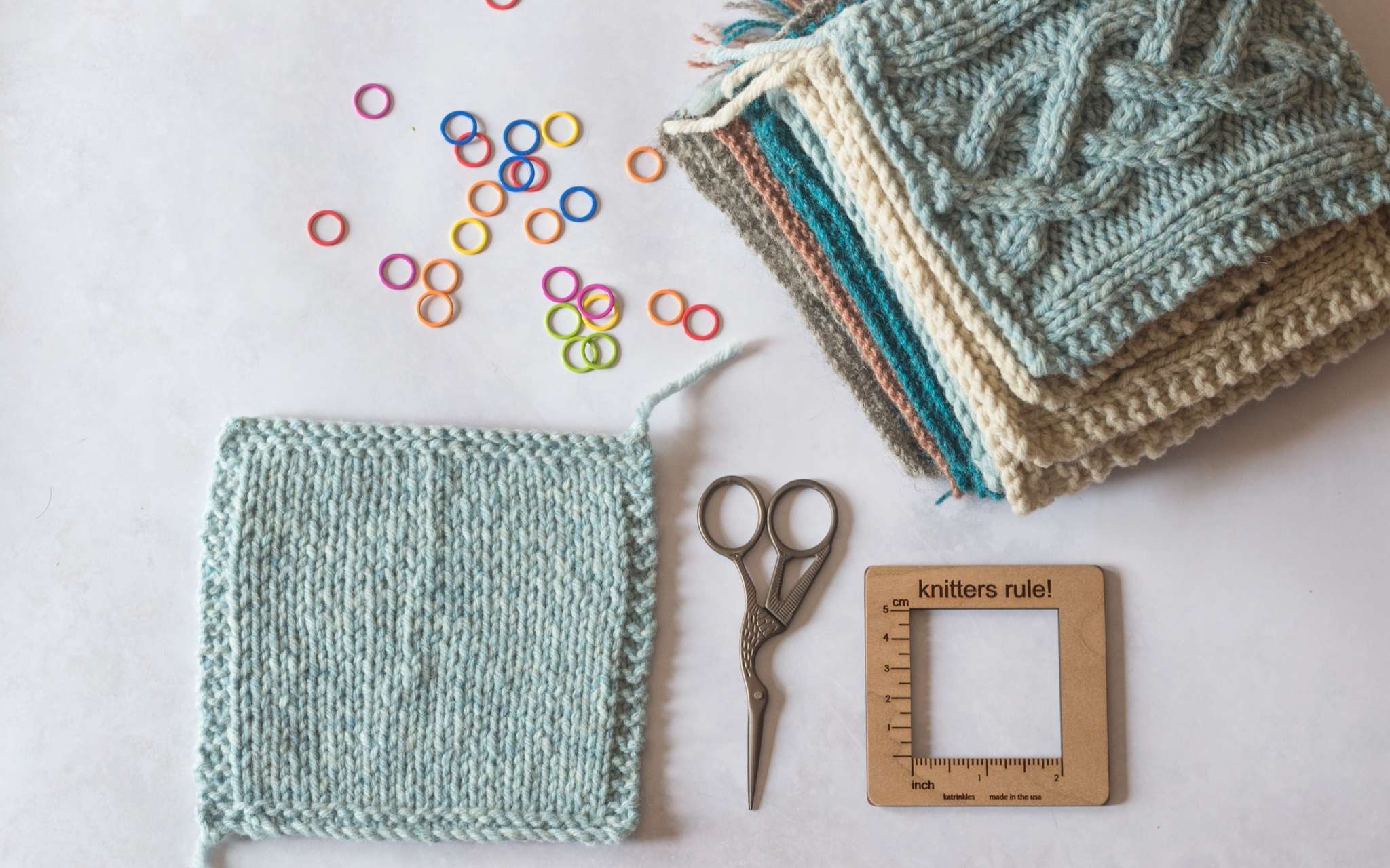 A blue stocking stitch swatch lies on a pale flat surface. Next to it is a small pair of scissors, a wooden gauge tool, coloured stitch markers and a pile of swatches.
