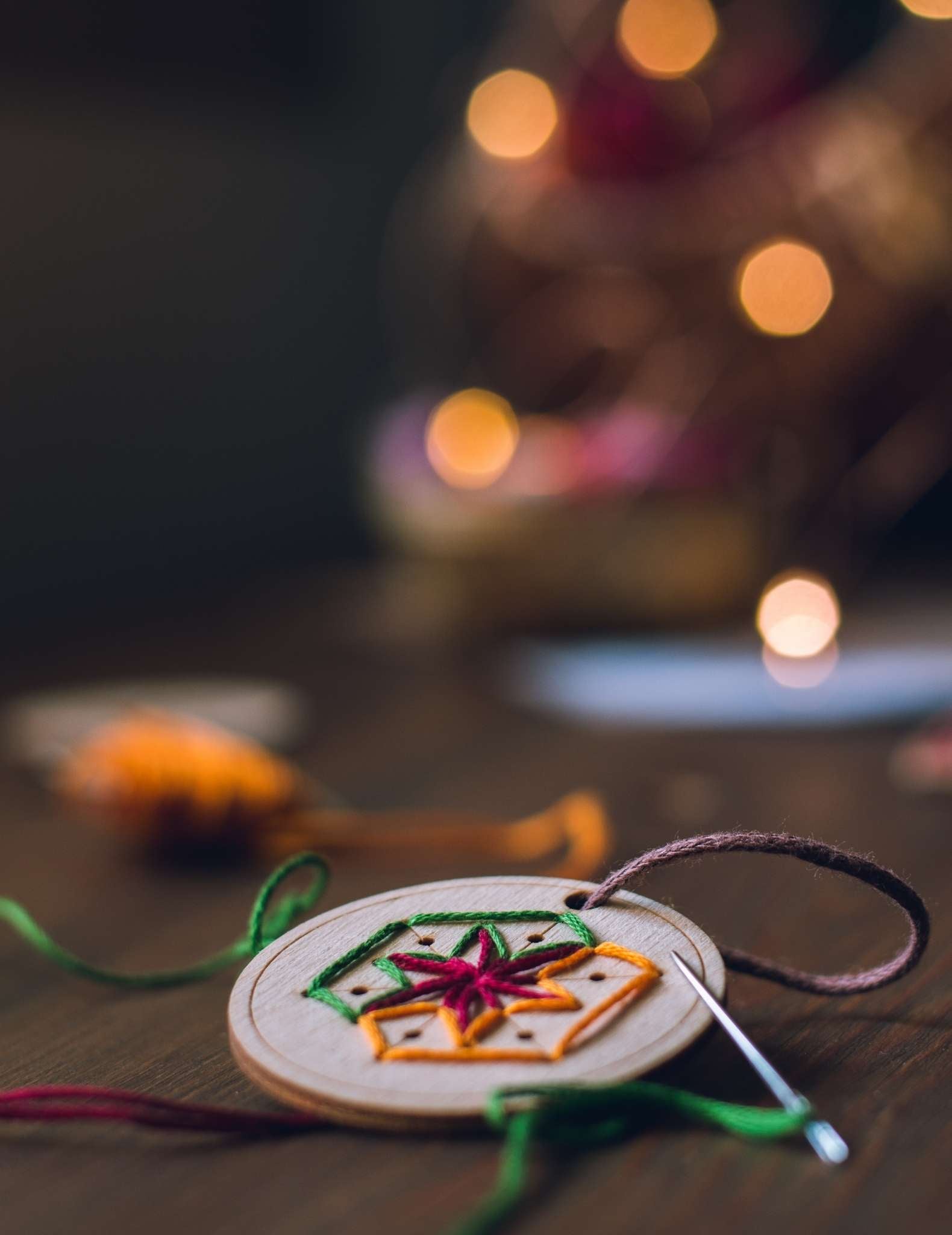 A circular wooden ornament is being hand stitched in a snowflake motif with brightly coloured threads. It lies on a table in front of a tree with fairy lights.