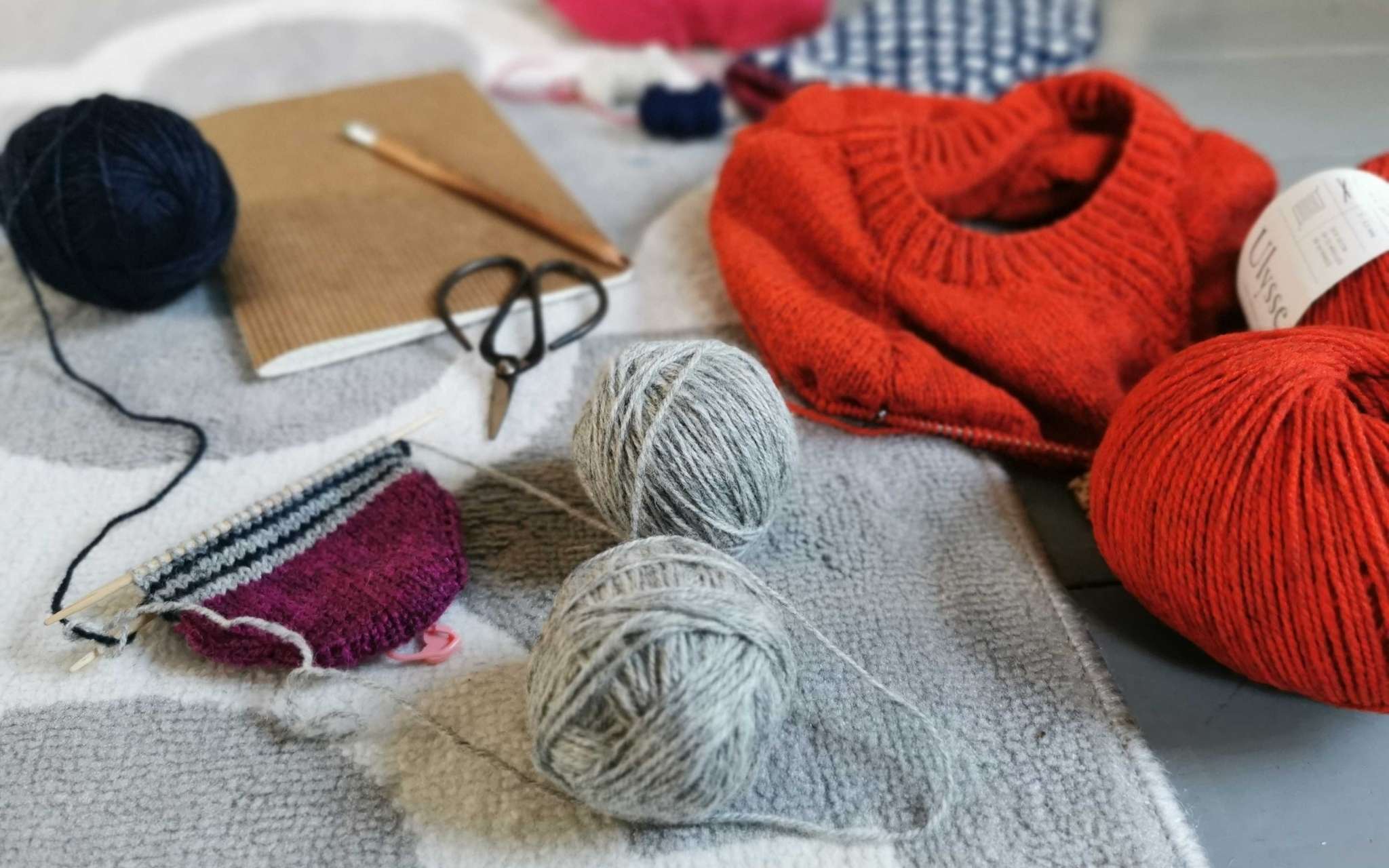 A pile of knitting projects on the needles including an orange sweater, striped socks next to a notebook and pencil.