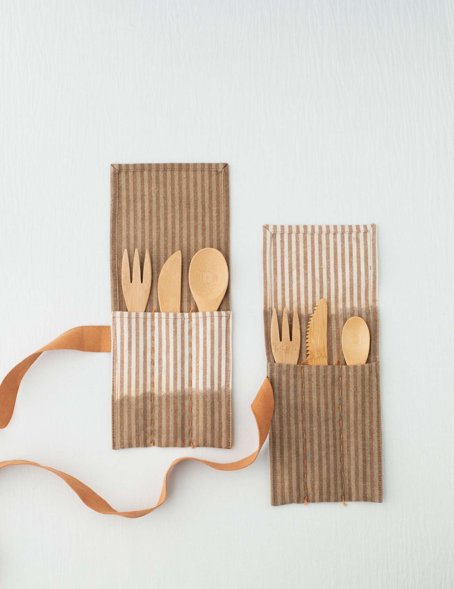Two brown and natural patterned utensil rolls laying flat on a white surface with ribbon closures hanging to the side. They are filled with a wooden fork, knife and spoon each.