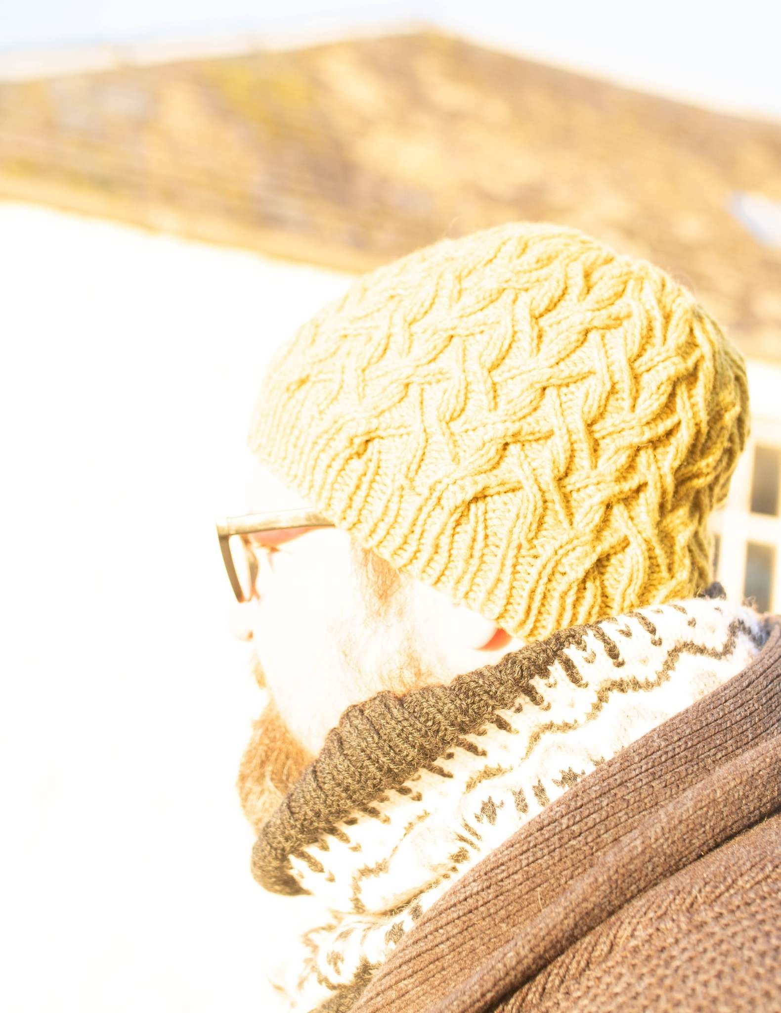 an over exposed photo of a white man with a beard and glasses, looking to the side, wearing a cabled beanie hat.