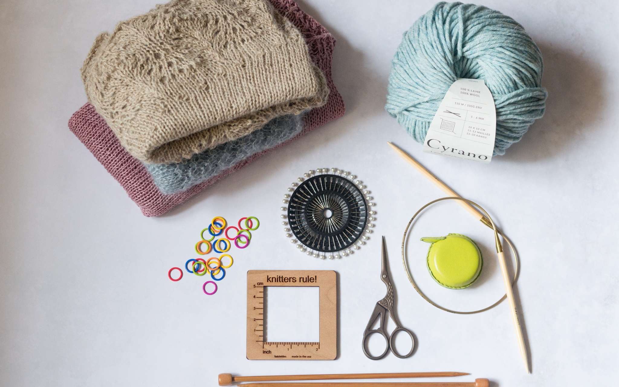 On a white flat surface lie a pile of knitted hats, a pale blue ball of yarn, straight and circular knitting needles, small scissors, a wooden square measuring tool and brightly coloured stitch markers.